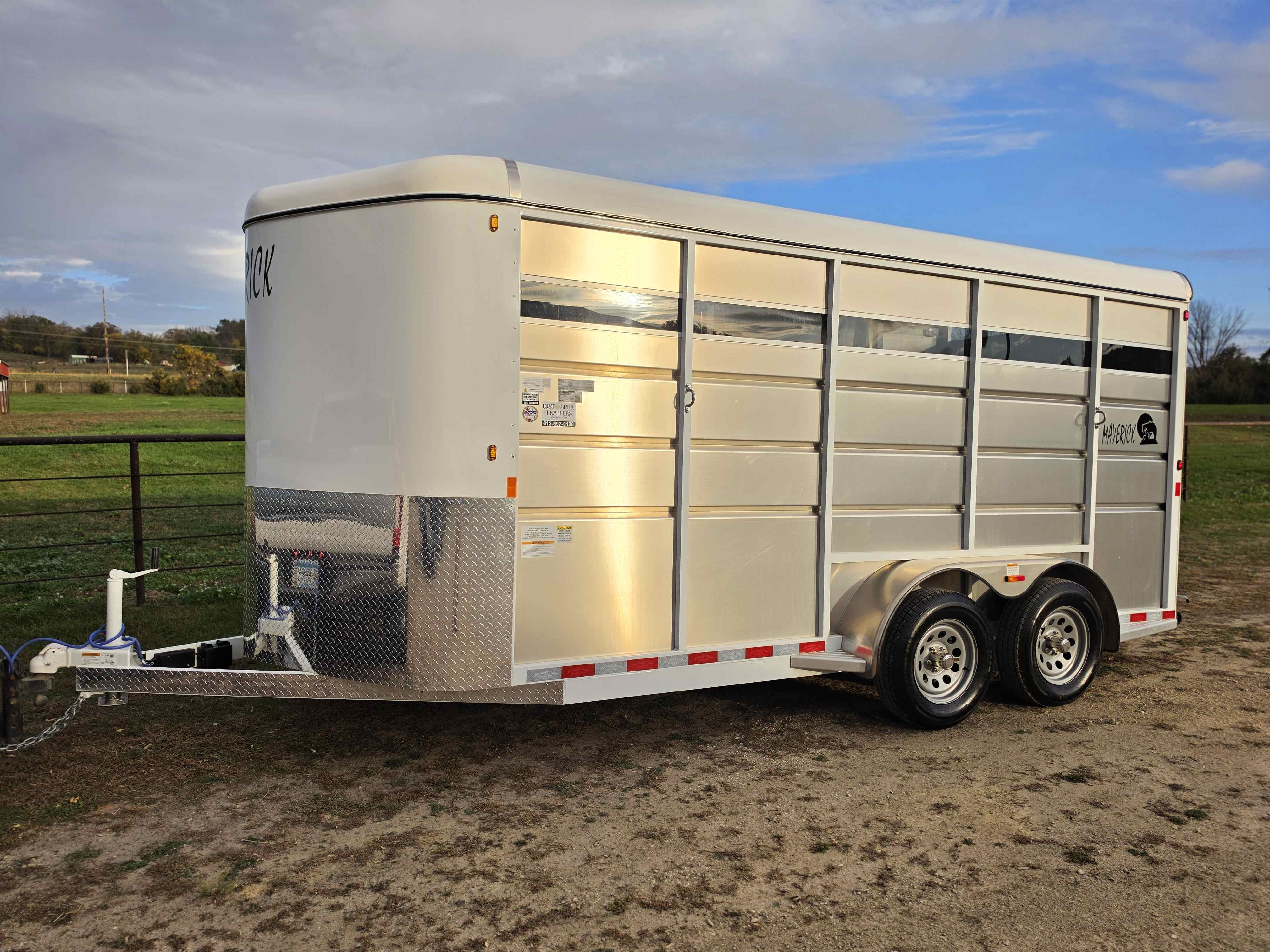 White and silver Maverick 3 horse stock combo trailer front-left view with diamond plate nose parked in a pasture