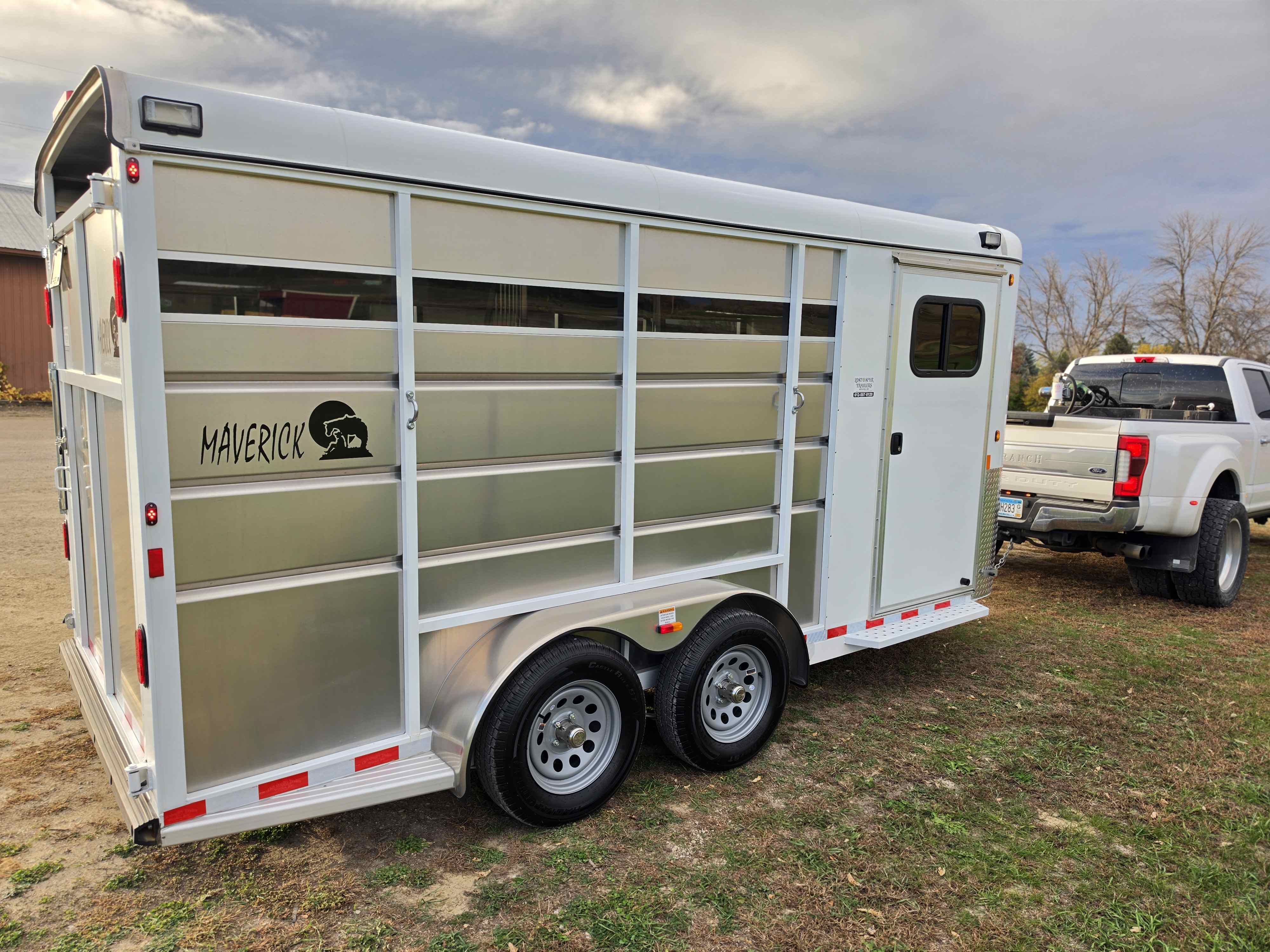 Maverick 3 horse stock combo trailer rear-left view with tandem axles and side tack room door beside a white dually truck