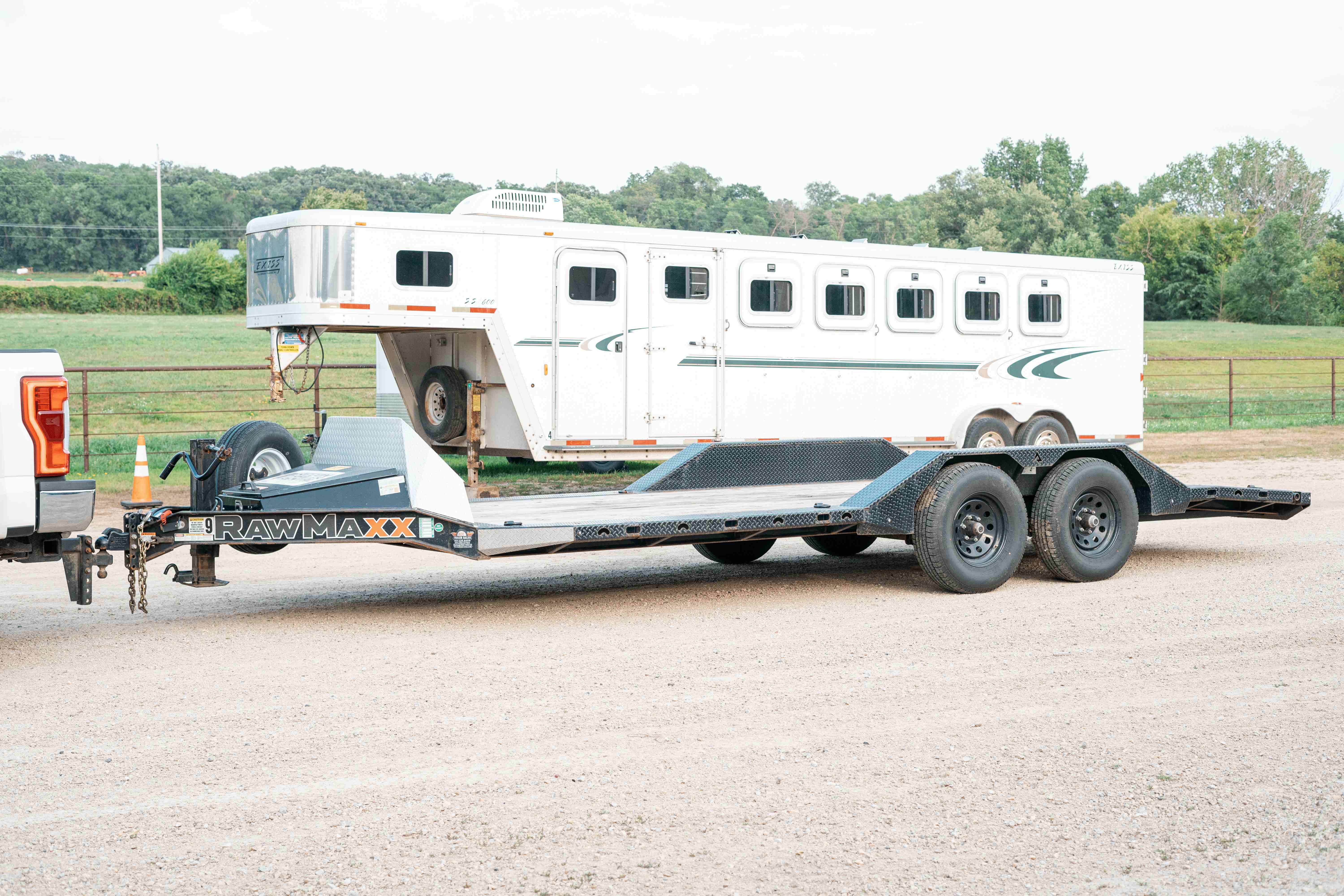 RawMaxx 20ft car trailer hitched to truck with empty low-deck platform, parked on gravel next to a horse trailer